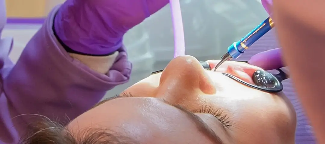A Carol trabalha na boca de uma paciente deitada, de olhos fechados.